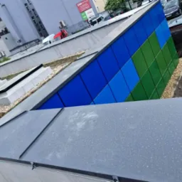 Nowoczesny dach z panelami w odcieniach niebieskiego i zieleni. Widoczne okna dachowe i fragment zielonego dachu. Tło stanowi zabudowa miejska.
