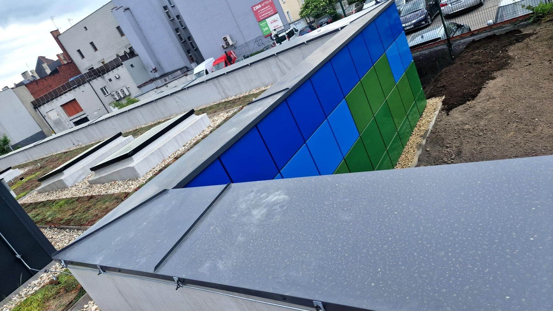 Nowoczesny dach z panelami w odcieniach niebieskiego i zieleni. Widoczne okna dachowe i fragment zielonego dachu. Tło stanowi zabudowa miejska.