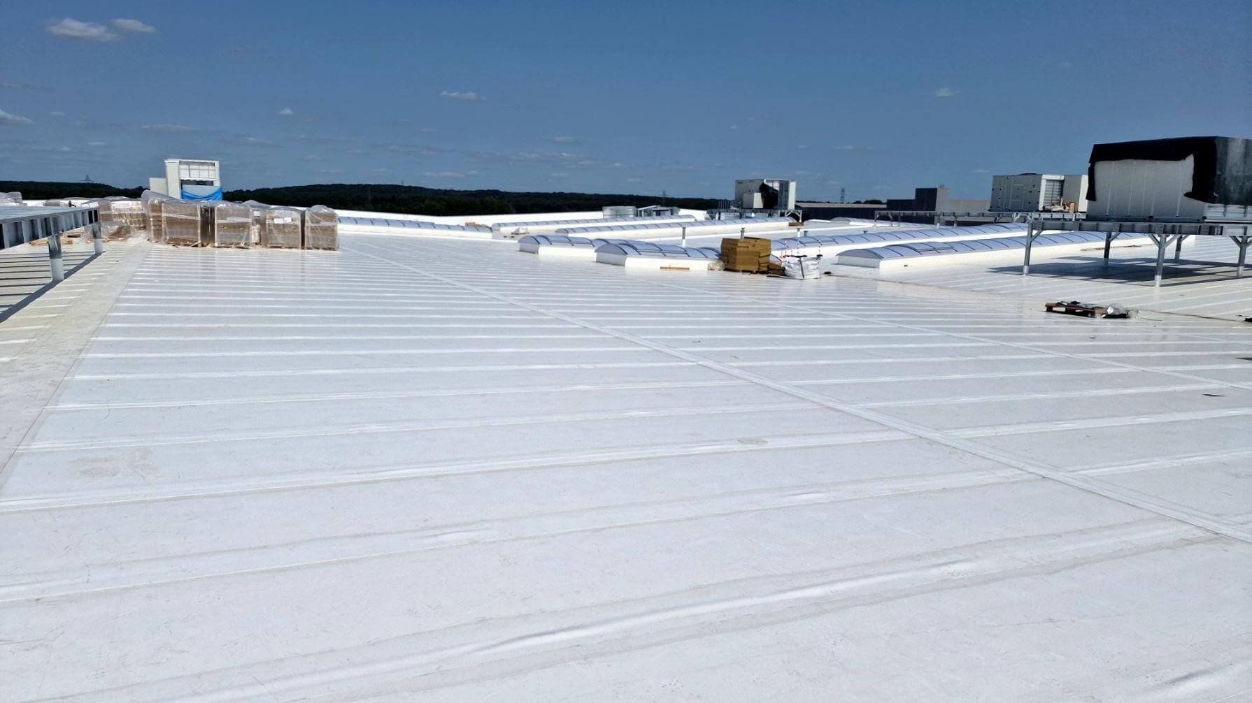 Biały dach przemysłowy z świetlikami i paletami materiałów budowlanych w tle, widok z góry w słoneczny dzień. Układ poziomy.