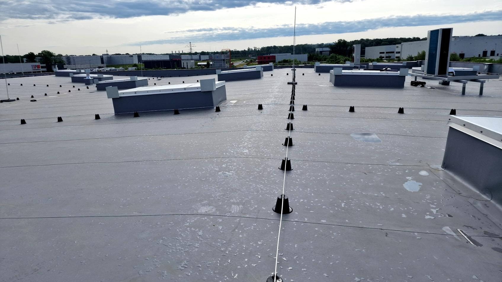 Szary dach płaski z instalacją odgromową i elementami wentylacyjnymi, widok z góry na tle budynków przemysłowych pod pochmurnym niebem.