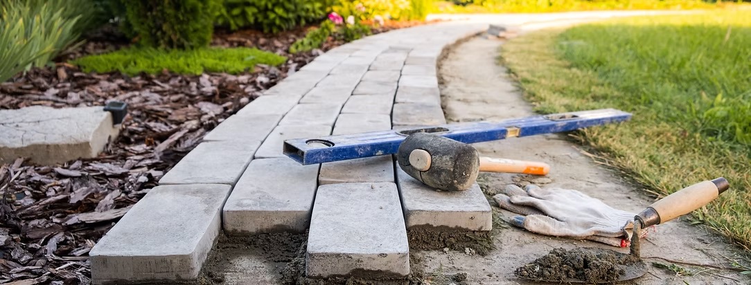 Szara kostka brukowa ułożona na ścieżce w ogrodzie, widoczny poziomica, gumowy młotek i rękawice robocze, co sugeruje etap prac brukarskich.