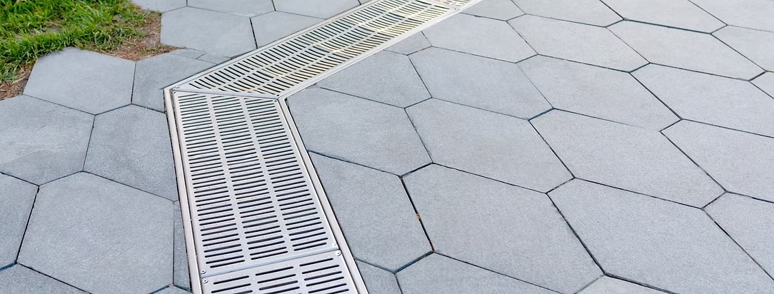 Szare, heksagonalne płyty chodnikowe z metalowym odpływem liniowym, widoczne z góry. Fragment trawy w lewym górnym rogu kadru. Geometryczny wzór.