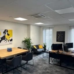 Nowoczesne biuro: dębowy stół konferencyjny z szarymi krzesłami, stanowiska pracy z monitorami, mapa Europy z logo firmy na ścianie, szara sofa z żółtą poduszką.