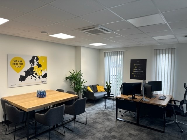 Nowoczesne biuro: dębowy stół konferencyjny z szarymi krzesłami, stanowiska pracy z monitorami, mapa Europy z logo firmy na ścianie, szara sofa z żółtą poduszką.