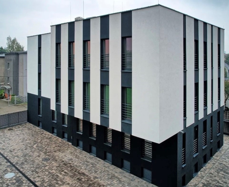 Nowoczesny budynek biurowy z biało-czarną elewacją, widoczny z perspektywy. Regularny układ okien z balustradami. Brukowany plac przed budynkiem. Minimalistyczny design.