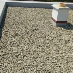 Płaski dach pokryty drobnymi kamieniami, z widocznym kominem w tle i szarym obrzeżem.