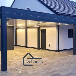 IwTaras - Nowoczesna, ciemna konstrukcja z oświetleniem LED, przylegająca do domu z ciemną dachówką. Widoczne drewniane elementy dekoracyjne i kostka brukowa.