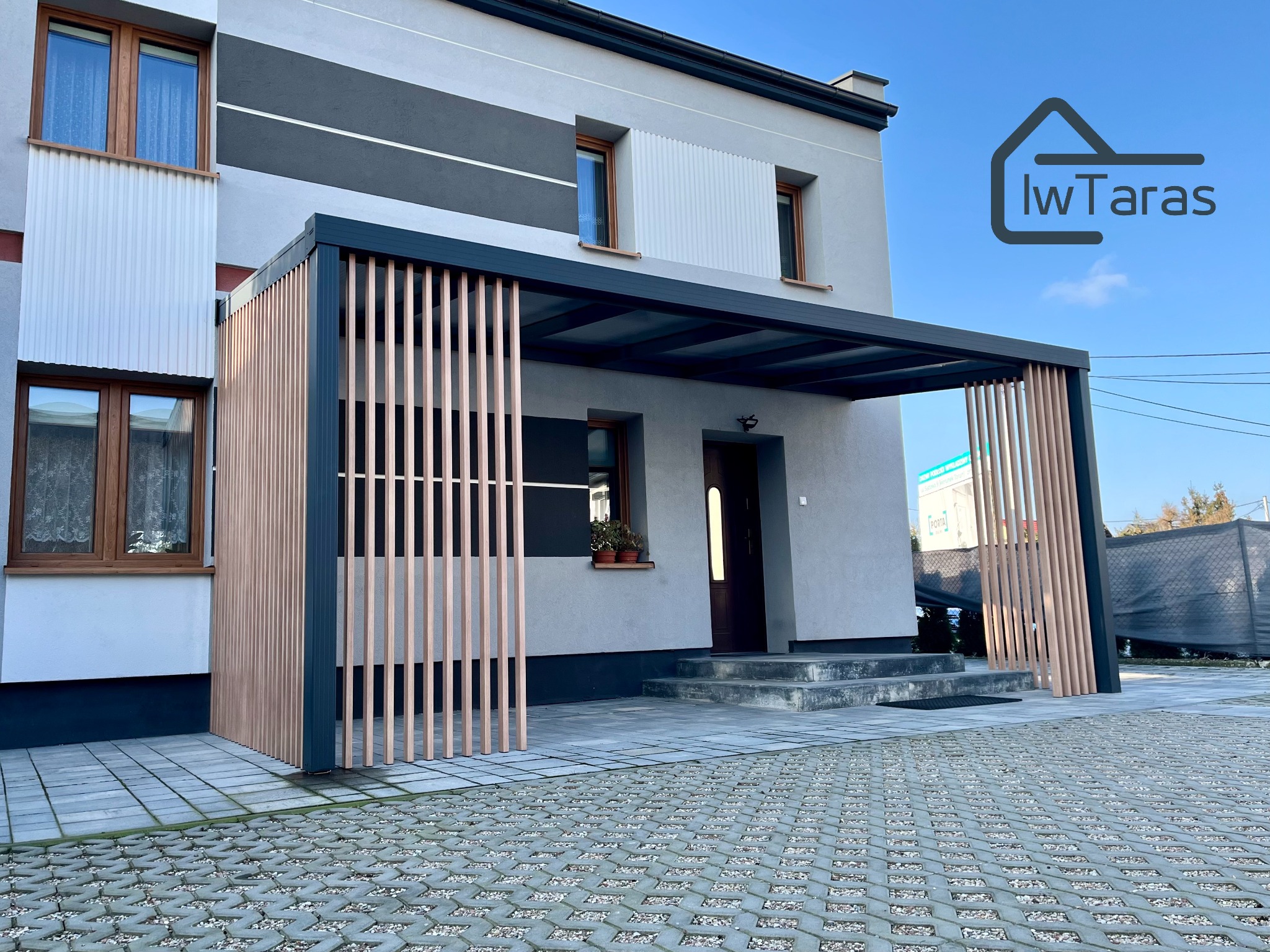 Nowoczesna pergola z drewnianymi lamelami i czarną ramą, przylegająca do budynku z szarą elewacją. Kostka brukowa na podłożu.