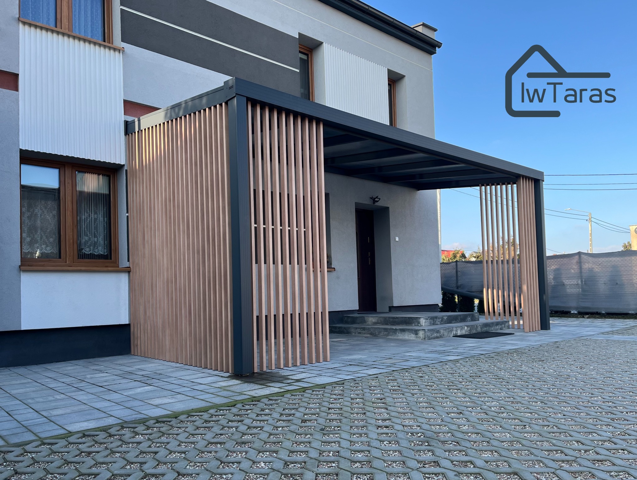 Nowoczesna pergola z drewnianymi lamelami przy wejściu do domu, z logo firmy IwTaras w prawym górnym rogu. Widok na szarą kostkę brukową i elewację budynku.