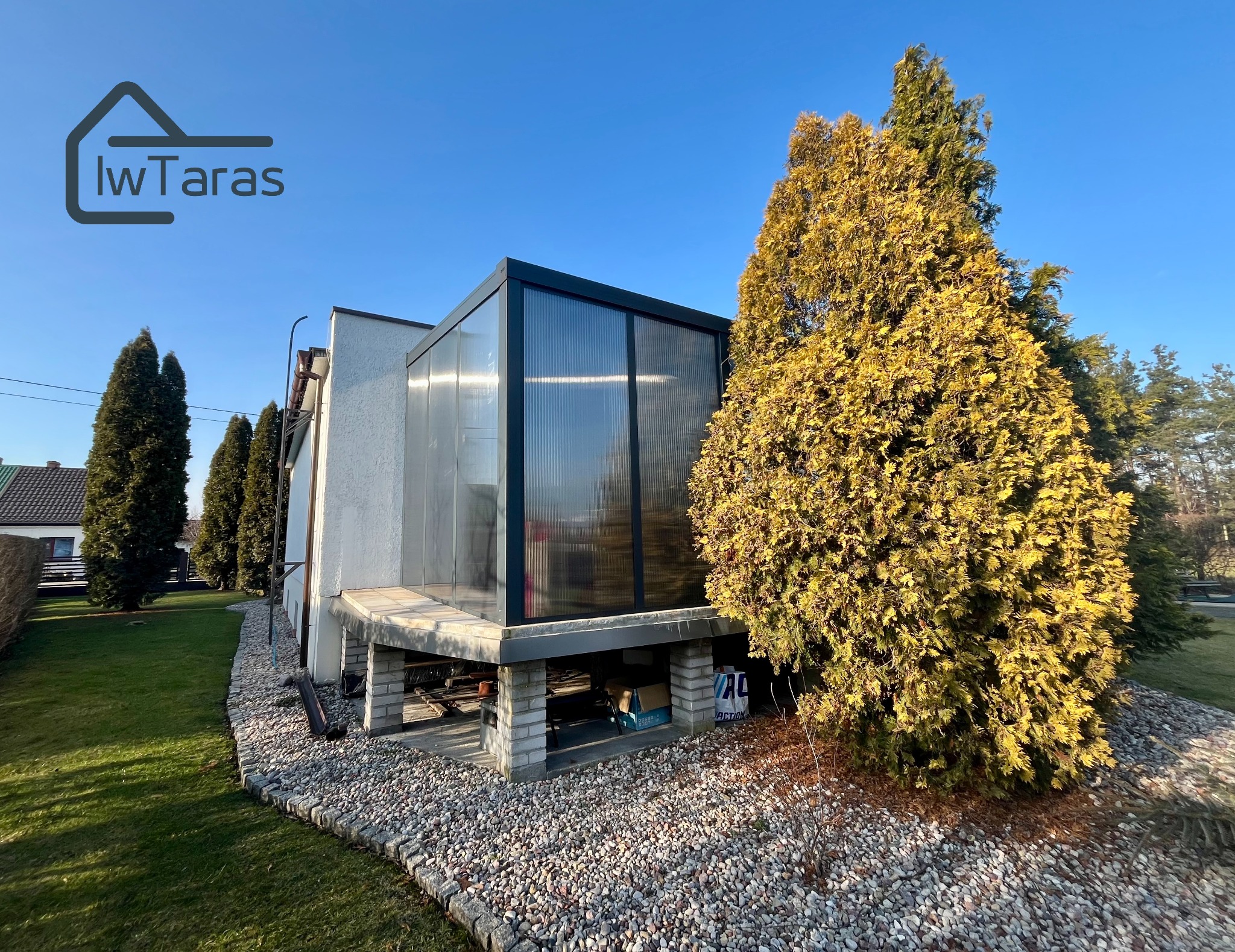 Nowoczesna zabudowa tarasu z przeszkleniami i czarną ramą, widoczna z boku, otoczona zielenią i kamieniami. Słoneczny dzień.