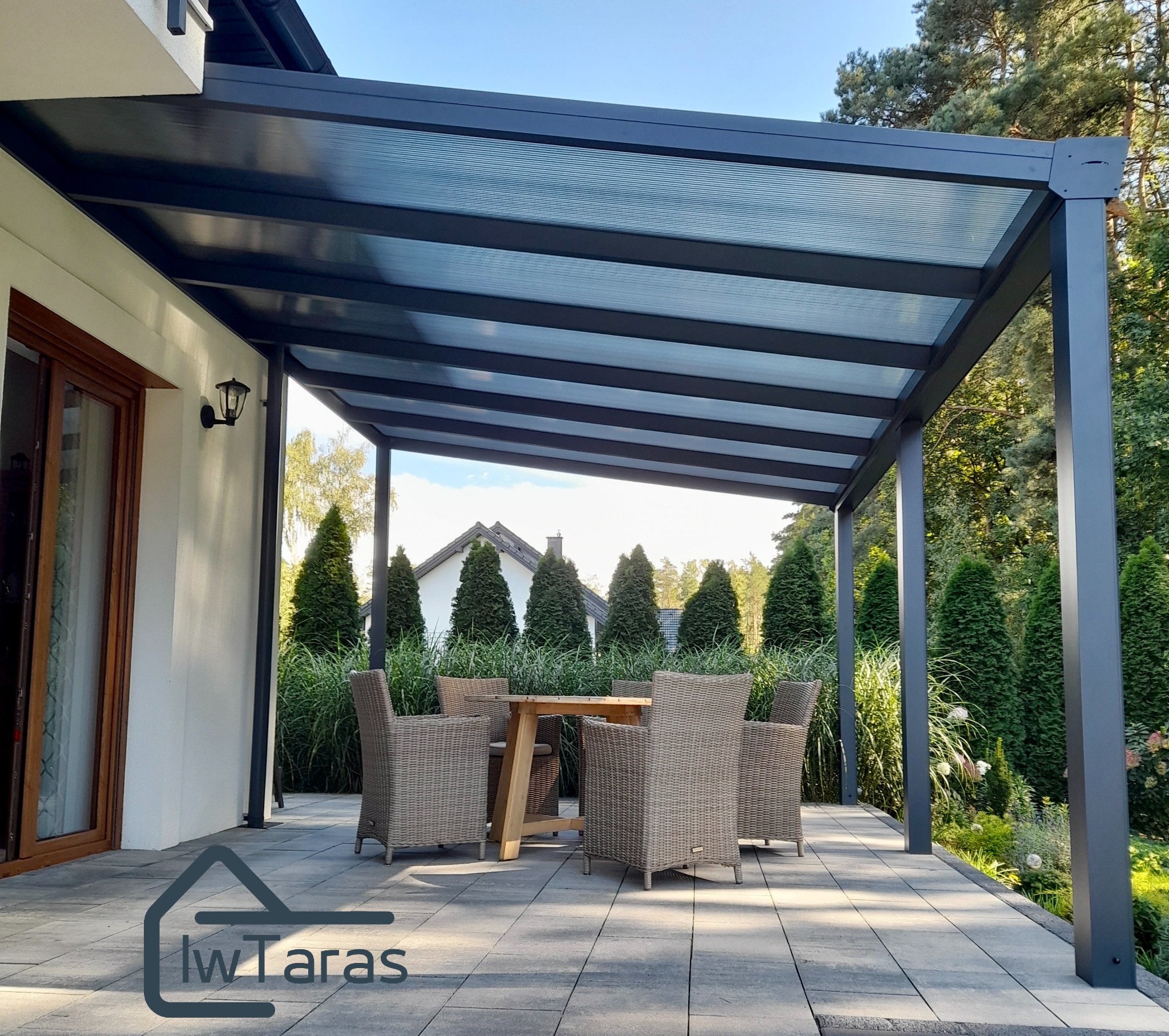 Nowoczesna, grafitowa pergola metalowa z przezroczystym zadaszeniem na tarasie. W tle zadbany ogród i dom. Na tarasie meble ogrodowe.