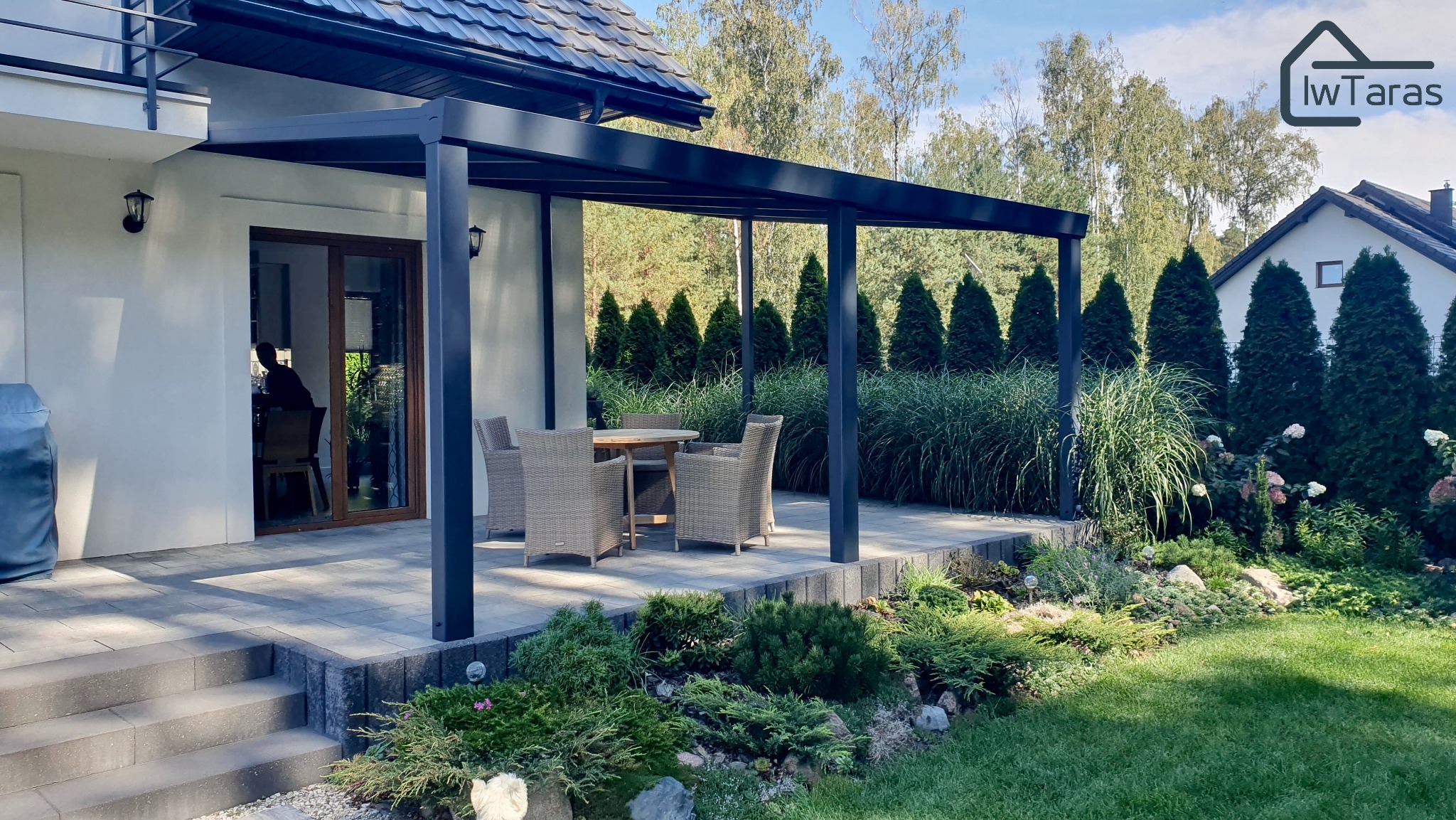 Nowoczesna, grafitowa pergola przy domu z tarasem. W tle rząd tuj i trawy ozdobne. Wygodne meble ogrodowe zapraszają do relaksu na świeżym powietrzu.