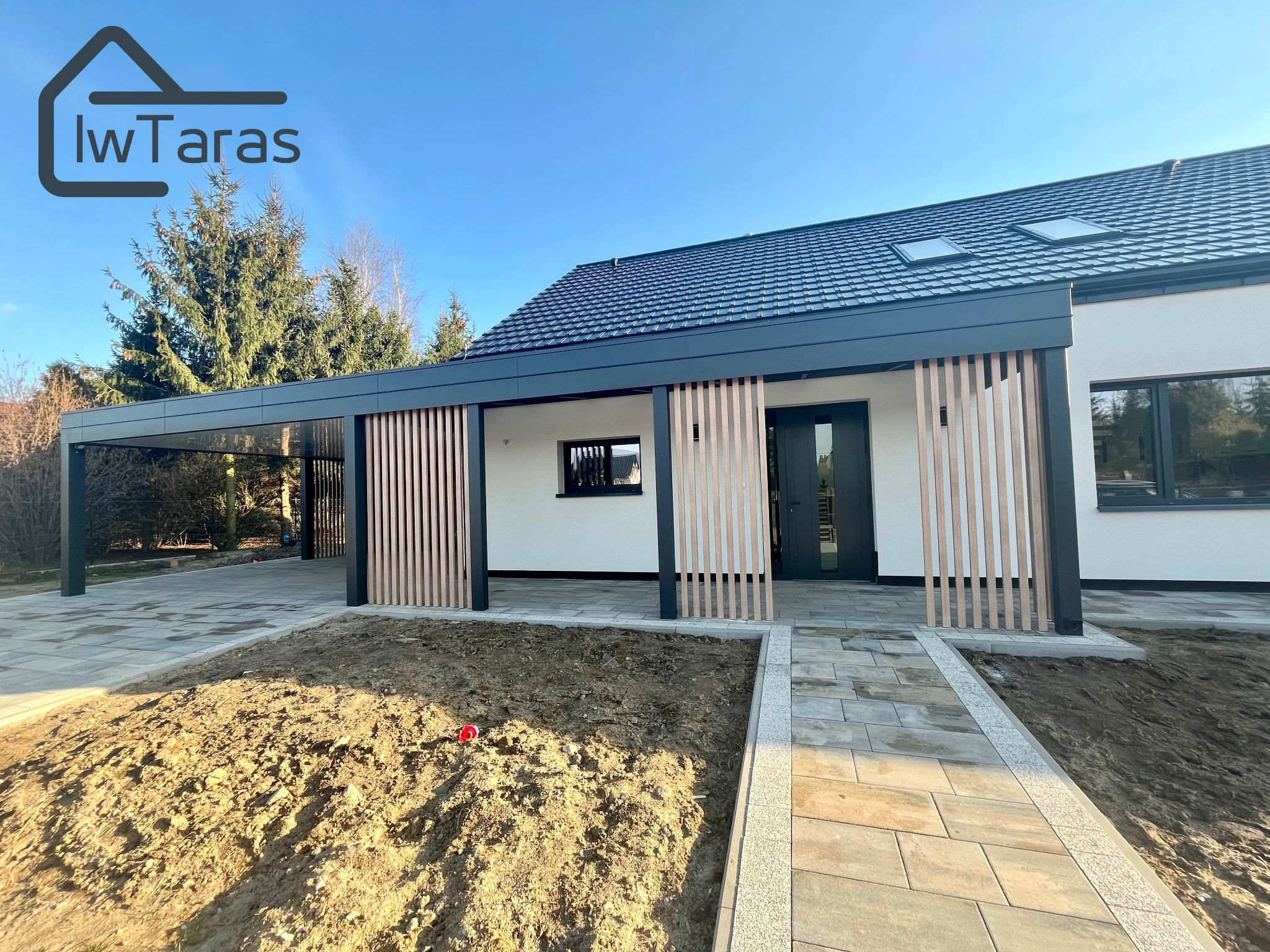 Nowoczesna pergola z metalową konstrukcją i drewnianymi lamelami, przylegająca do domu z ciemną dachówką. Ułożona kostka brukowa, ziemia przygotowana pod nasadzenia.