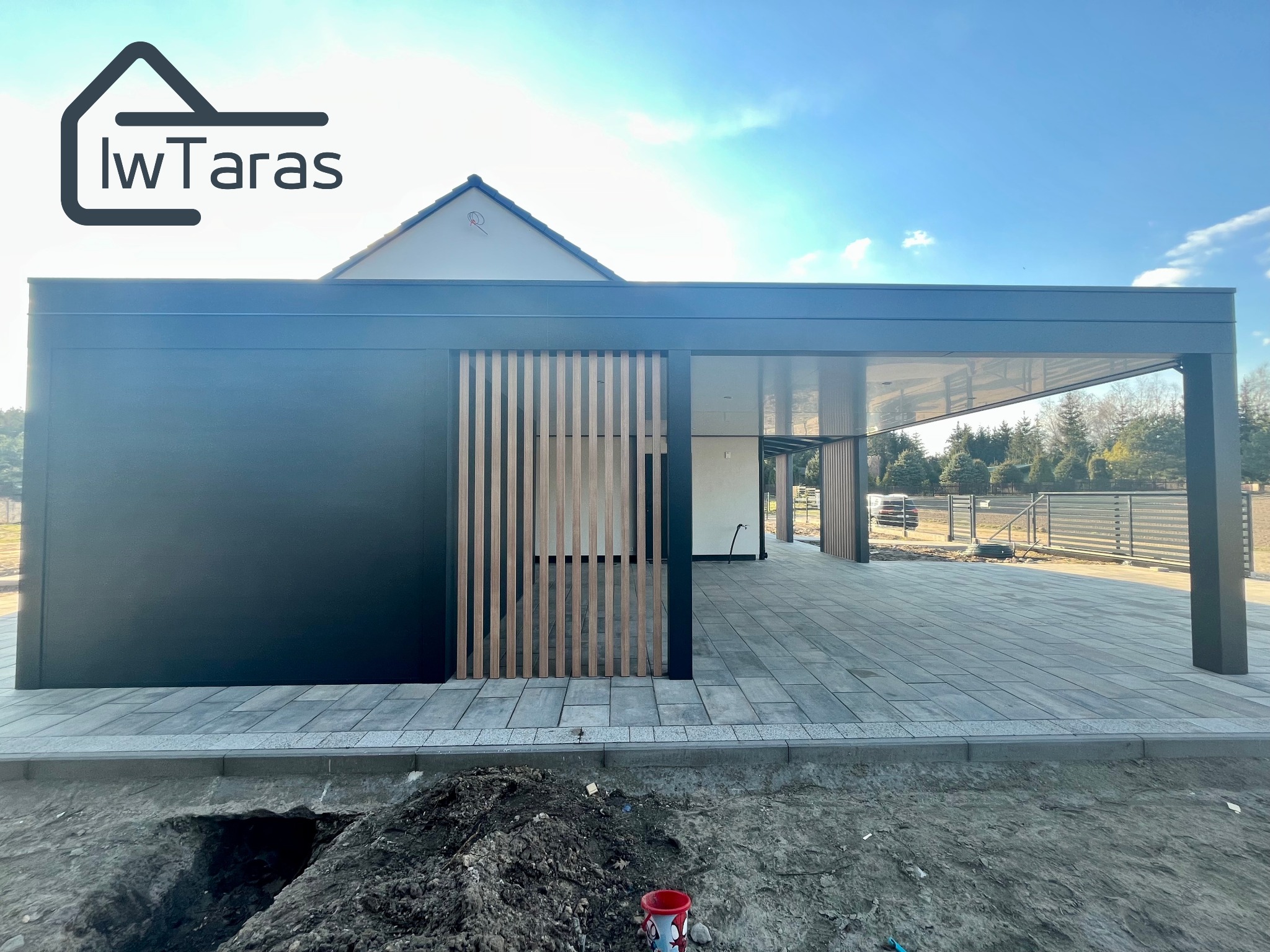 Nowoczesna, czarna pergola z drewnianymi lamelami, częściowo zabudowana, z widocznym fragmentem domu w tle i wybrukowanym tarasem. Widoczne wykończenia.