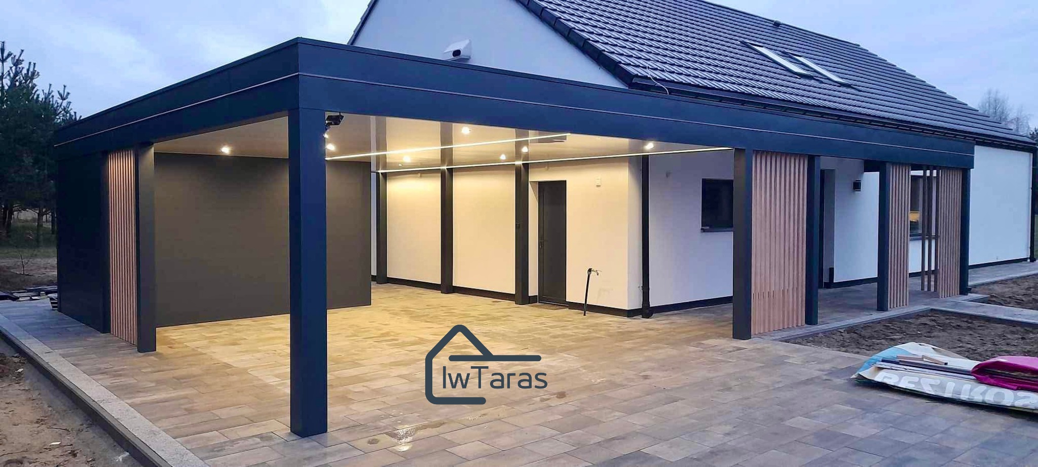 Nowoczesna, ciemna konstrukcja z oświetleniem LED, przylegająca do domu z ciemną dachówką. Widoczne drewniane elementy dekoracyjne i kostka brukowa.