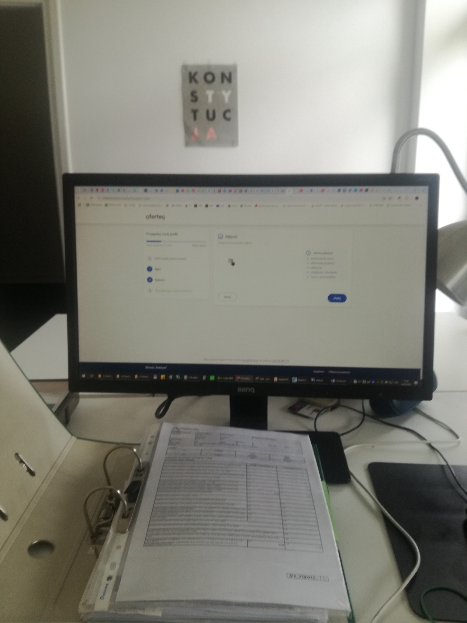 Monitor z otwartą stroną Oferteo, obok segregator z dokumentami. Widoczne biurko, lampka i plakat z napisem 'Konstytucja'.