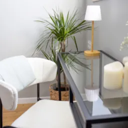 Jasne wnętrze: krzesło z pledem, szklany stolik z dekoracjami (świece, kwiaty), roślina doniczkowa. Stonowana kolorystyka, minimalistyczny styl.