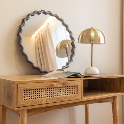 DAVI HOUSE SPÓŁKA Z OGRANICZONĄ ODPOWIEDZIALNOŚCIĄ - Drewniana toaletka z rattanową szufladą, okrągłe lustro z falistą ramą i złota lampka na blacie. Otwarta książka. Beżowe ściany i zasłony w tle.