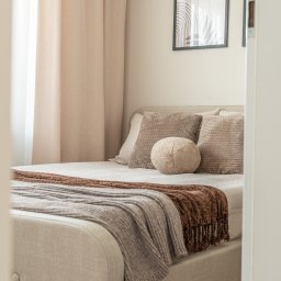 DAVI HOUSE SPÓŁKA Z OGRANICZONĄ ODPOWIEDZIALNOŚCIĄ - Beżowa sypialnia z dekoracyjnymi poduszkami i pledem, widziana zza framugi drzwi. Stonowana kolorystyka, minimalistyczny wystrój, rama obrazu na ścianie.