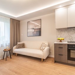 DAVI HOUSE SPÓŁKA Z OGRANICZONĄ ODPOWIEDZIALNOŚCIĄ - Jasne wnętrze: kremowa sofa z poduszkami, stolik z tacą, okno z firanką i zasłoną, zabudowa kuchenna z marmurowym blatem i piekarnikiem. Minimalistyczny design.