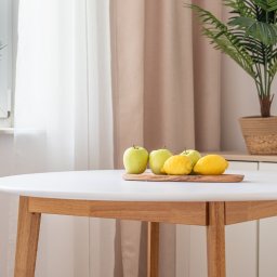 DAVI HOUSE SPÓŁKA Z OGRANICZONĄ ODPOWIEDZIALNOŚCIĄ - Jasne wnętrze: okrągły stół z owocami (jabłka, cytryny) na drewnianej desce. W tle roślina doniczkowa, okno z zasłonami. Minimalistyczny wystrój, naturalne światło.