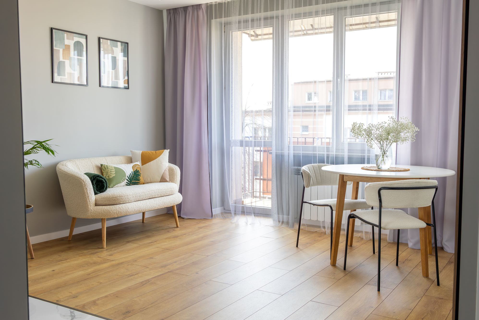 Jasny salon z drewnianą podłogą, kremową sofą z poduszkami, okrągłym stołem i krzesłami. Na stole wazon z gipsówką. Ściany w odcieniach szarości.