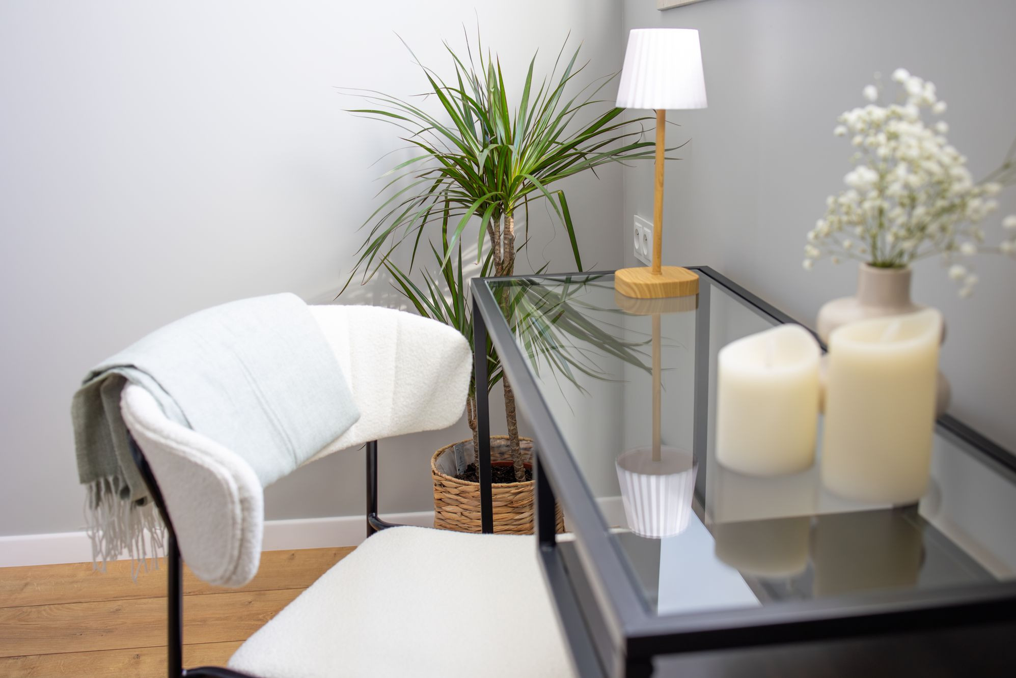 Jasne wnętrze: krzesło z pledem, szklany stolik z dekoracjami (świece, kwiaty), roślina doniczkowa. Stonowana kolorystyka, minimalistyczny styl.