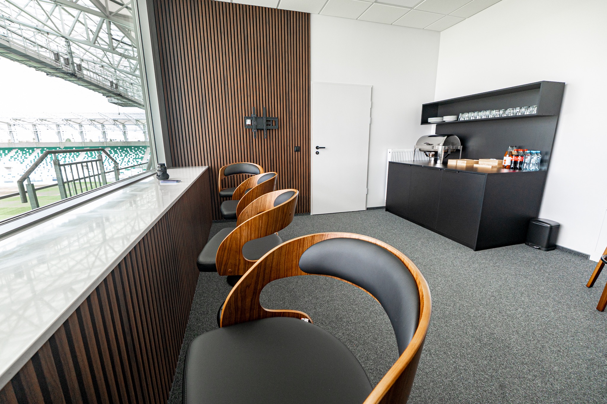 Nowoczesna loża VIP na stadionie z drewnianymi panelami, krzesłami o unikalnym designie i widokiem na boisko przez panoramiczne okno.