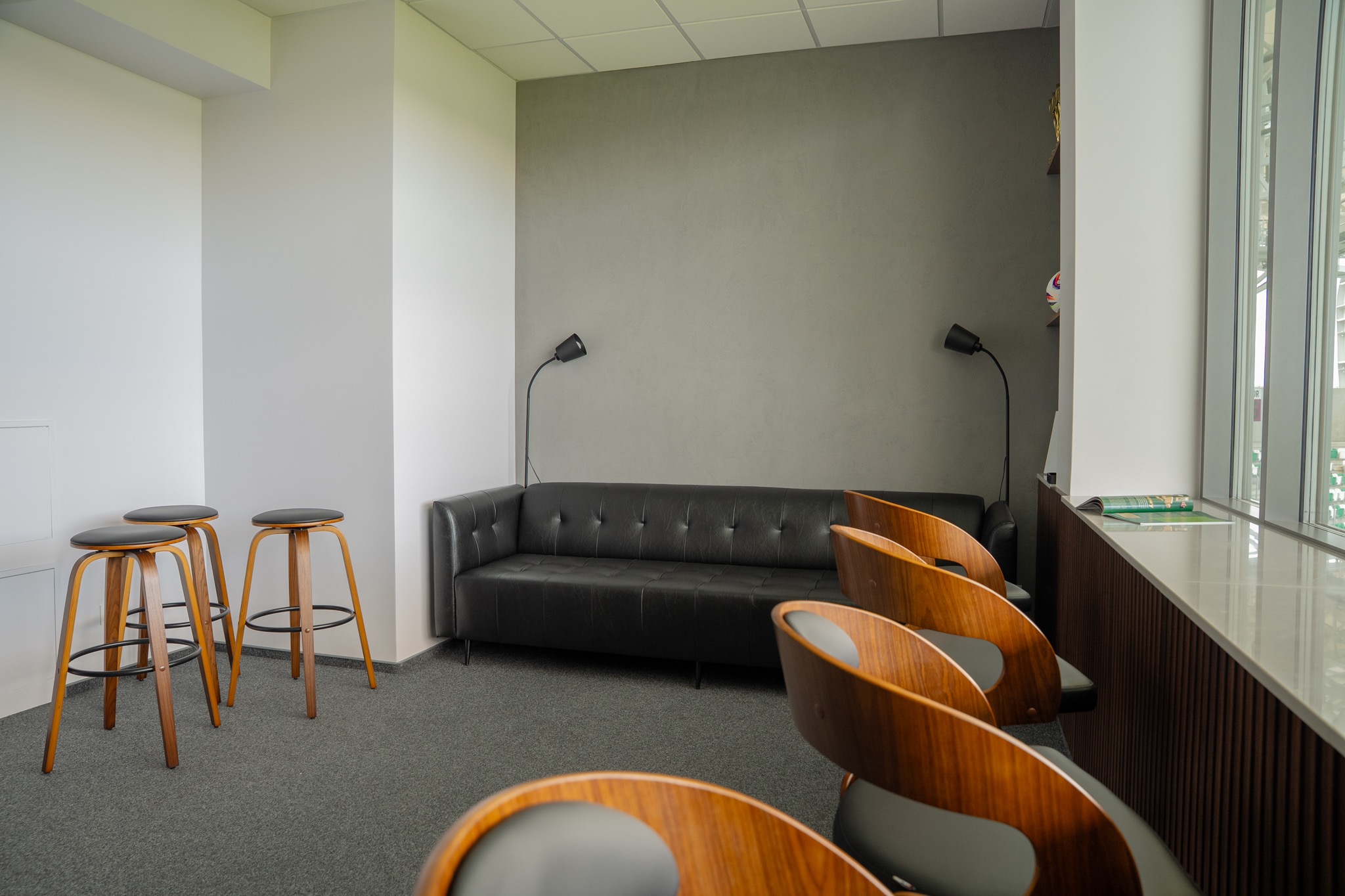 Nowoczesne wnętrze biurowe z czarną sofą, drewnianymi krzesłami barowymi i lampami podłogowymi. Stonowana kolorystyka, minimalistyczny design.