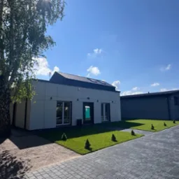 Nowoczesny budynek usługowy z szarą elewacją i zielonym trawnikiem w Pruszkowie. Widoczny fragment parkingu z kostki brukowej i drzewo liściaste.