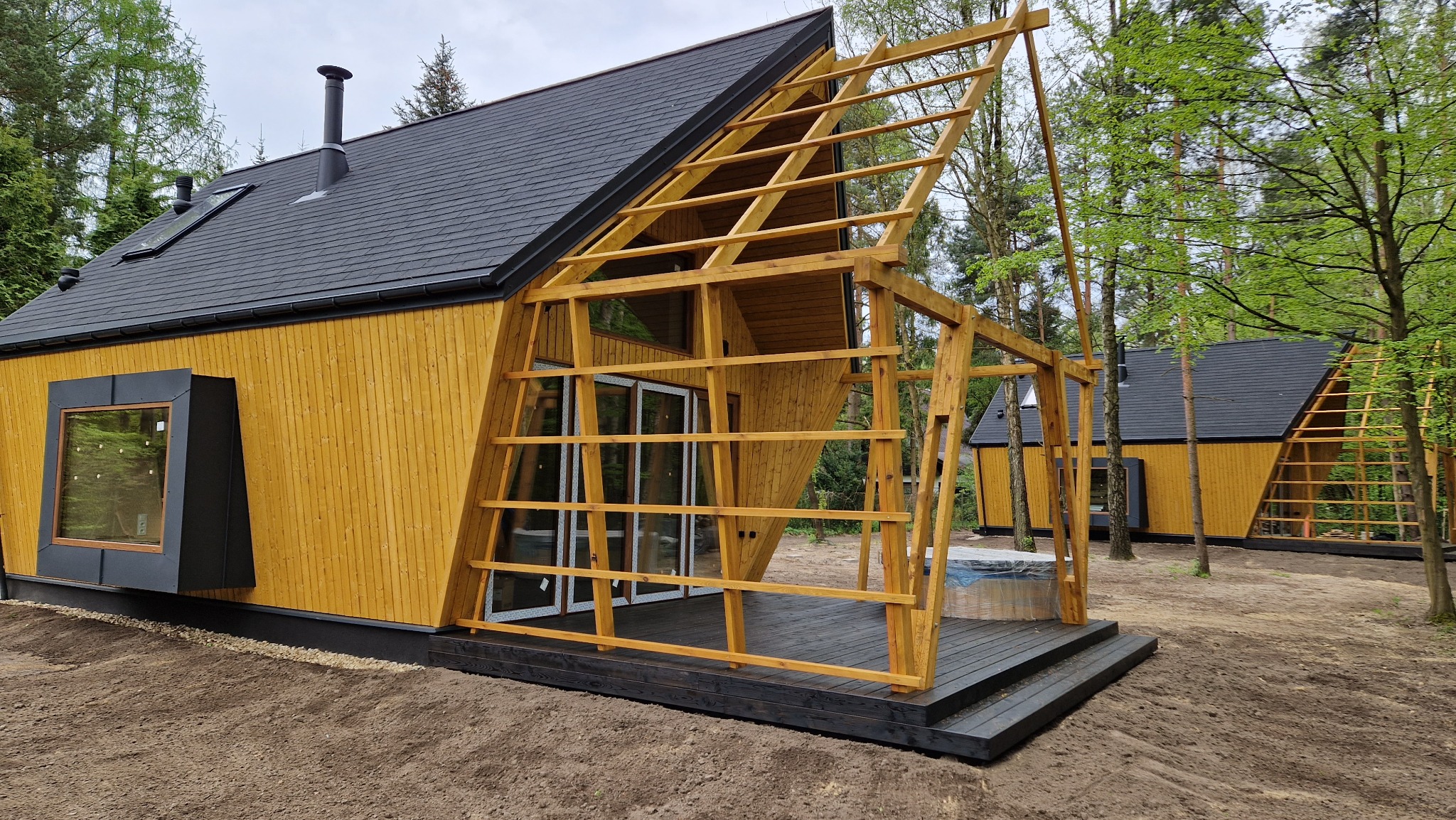 Żółty dom z drewnianą konstrukcją dachu i tarasu w lesie. Charakterystyczne okno w kształcie kostki. Częściowo zadaszony taras. Drugi podobny dom w tle.