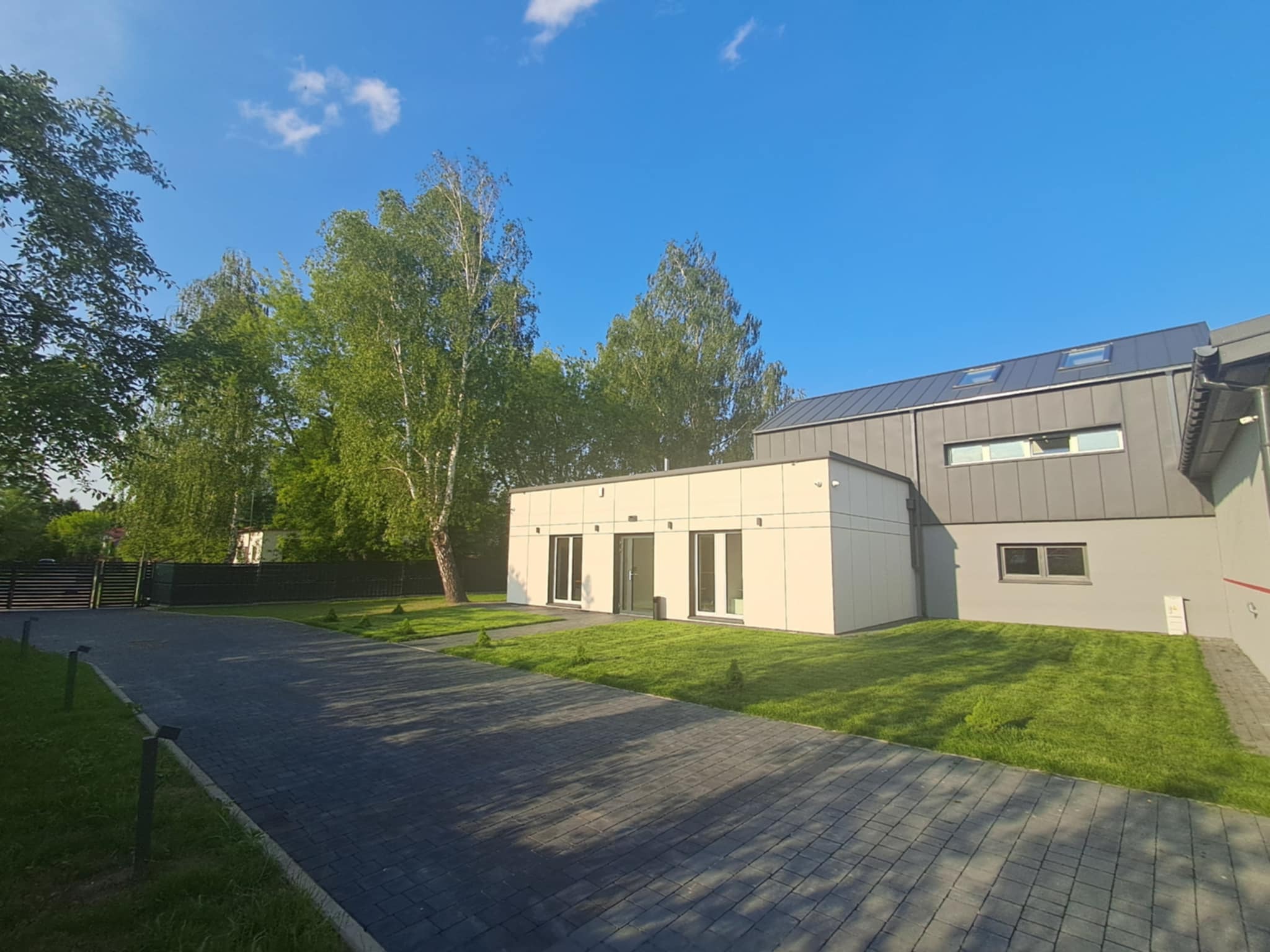 Nowoczesny budynek biurowy z grafitowym dachem i jasną elewacją, otoczony zielonym trawnikiem i brukowaną ścieżką w słoneczny dzień. Widoczne drzewa w tle.