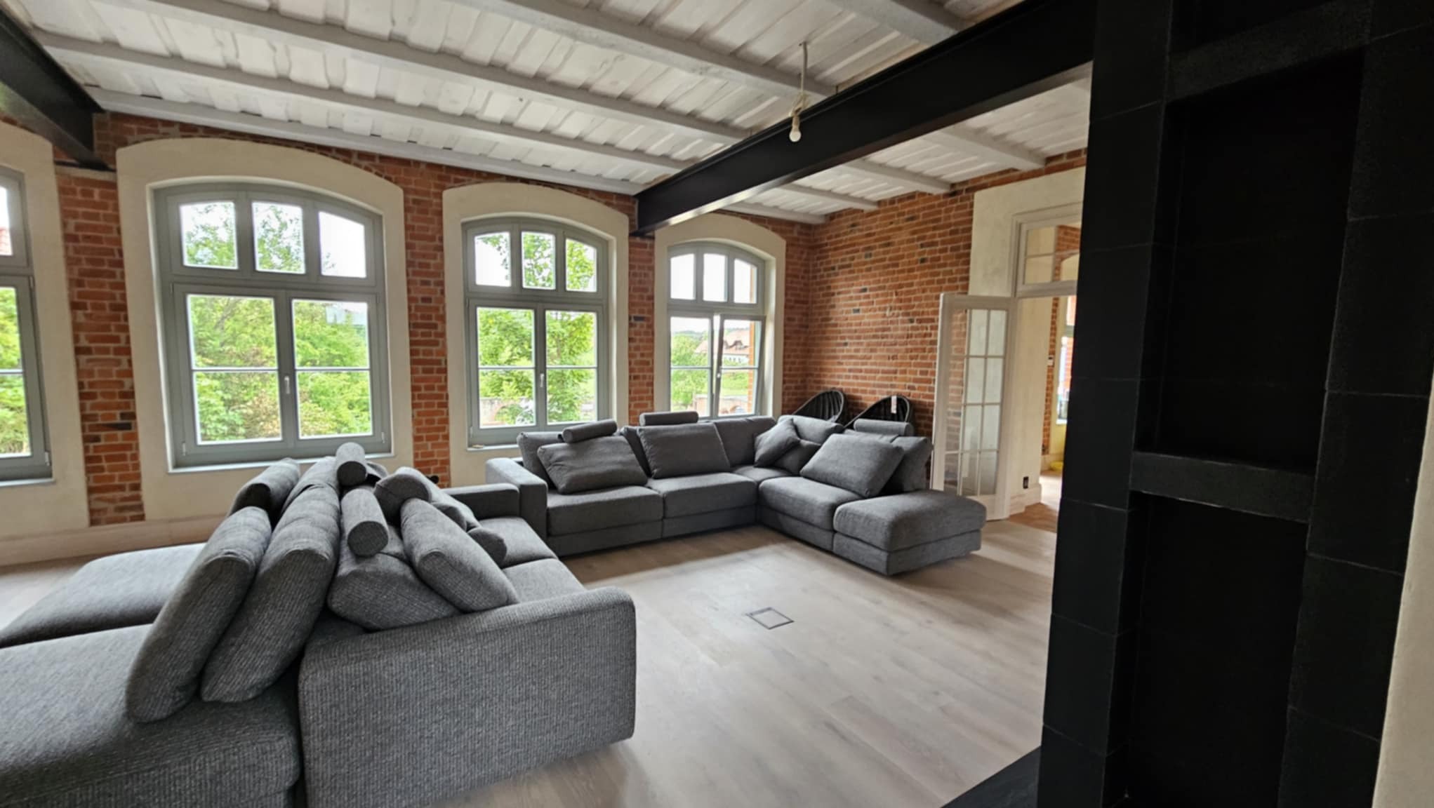 Loftowe wnętrze z ceglanymi ścianami, szarą sofą narożną i dużymi oknami z widokiem na zieleń. Biały sufit z czarnymi belkami. Parkiet w jasnym odcieniu drewna.