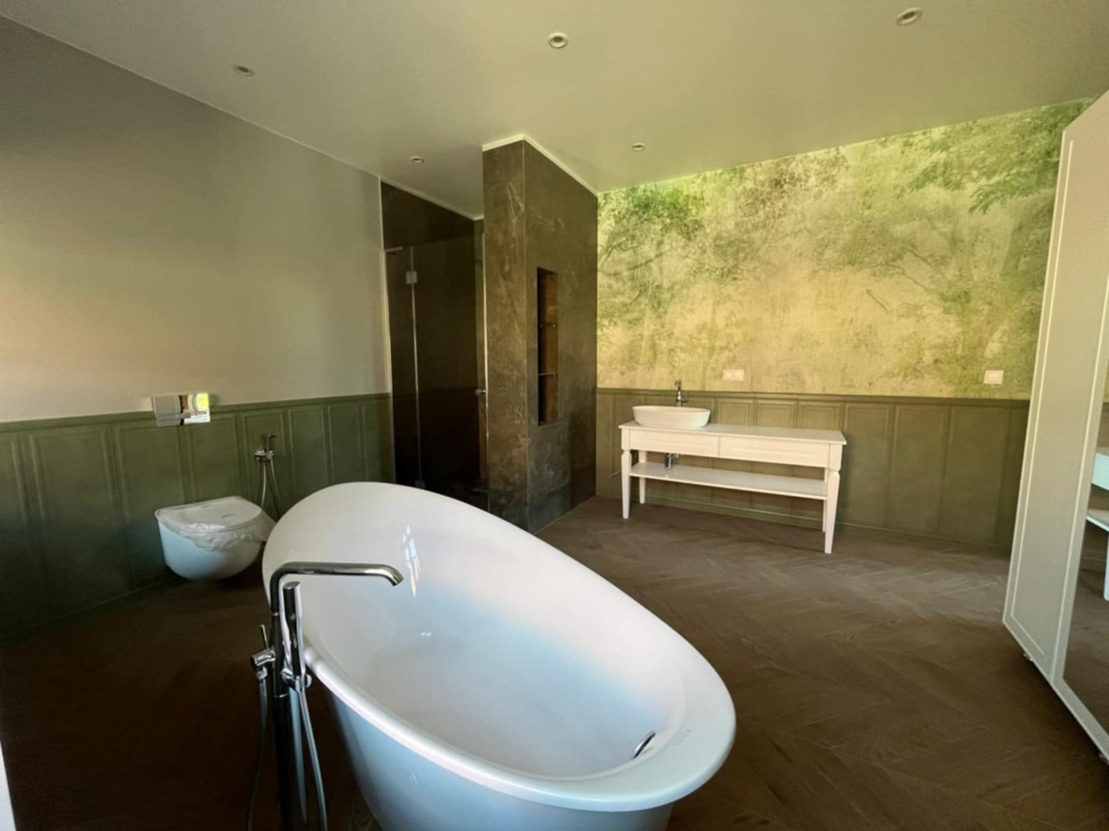 Elegancka łazienka z wolnostojącą wanną, zielonymi panelami ściennymi i fototapetą z motywem leśnym. Widoczna kabina prysznicowa i umywalka na białej konsoli.