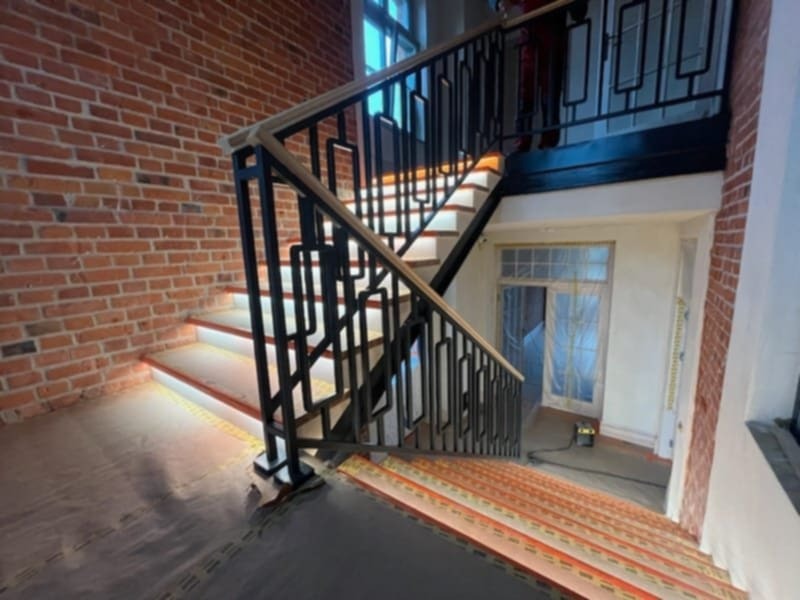 Klatka schodowa z oświetleniem LED pod stopniami, ceglana ściana, czarna metalowa balustrada o geometrycznym wzorze i osłonięte folią drzwi w tle.