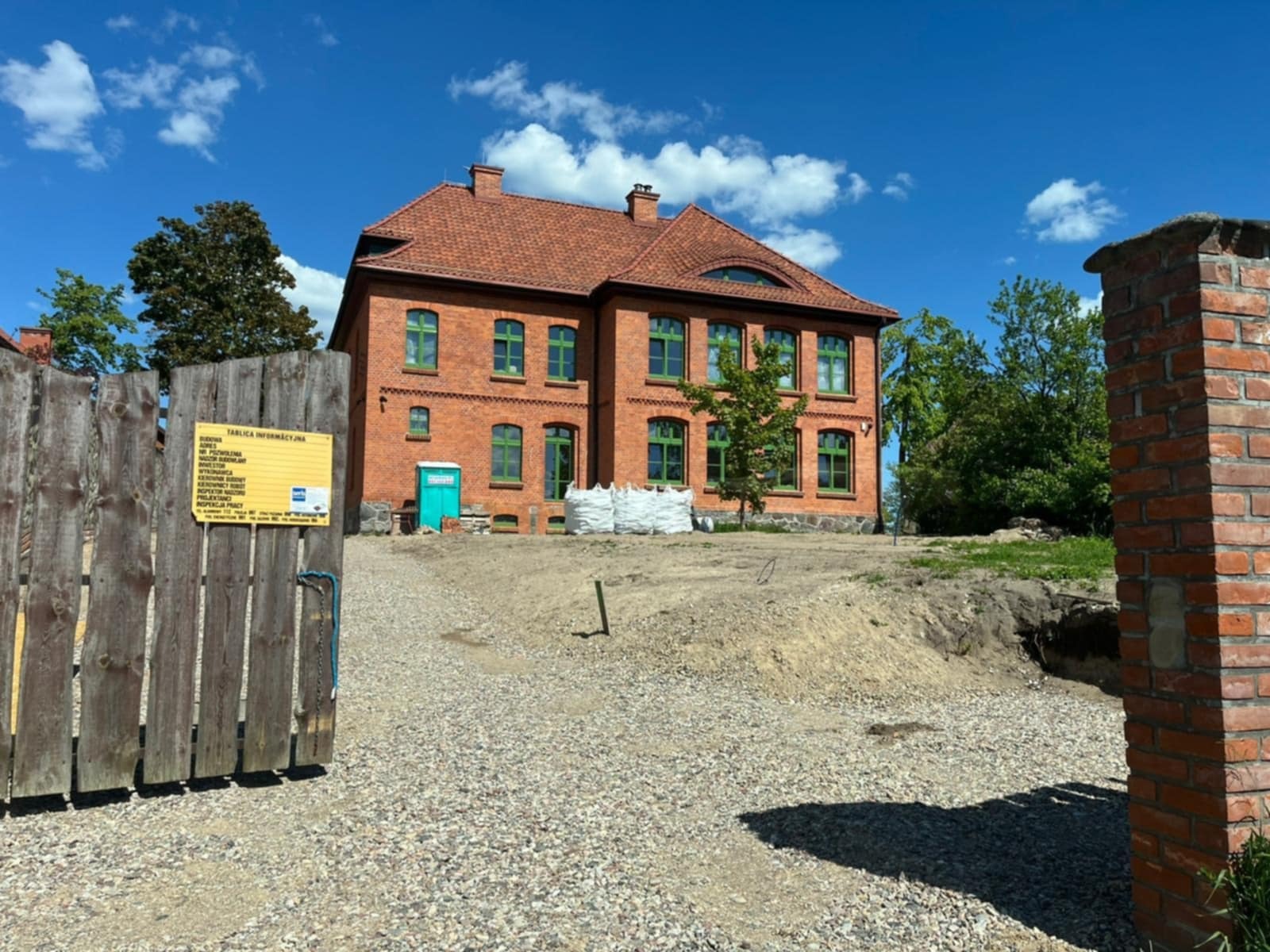 Ceglany budynek z zielonymi oknami i nową elewacją, widoczny zza drewnianego ogrodzenia z tablicą informacyjną, w słoneczny dzień.
