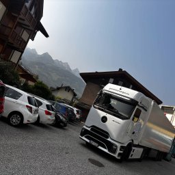 GENOMAR SP. Z O.O. - Biała ciężarówka Actros L z logo genomarcargo.eu na tle gór i parkingu z samochodami osobowymi. Ujęcie z dołu, pochylone w lewo.