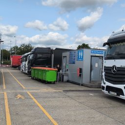 GENOMAR SP. Z O.O. - Parking dla ciężarówek z białymi Mercedesami Actros L firmy genomarcargo.eu. Na parkingu stoją zielone kontenery na śmieci. W tle widoczny budynek z toaletami.