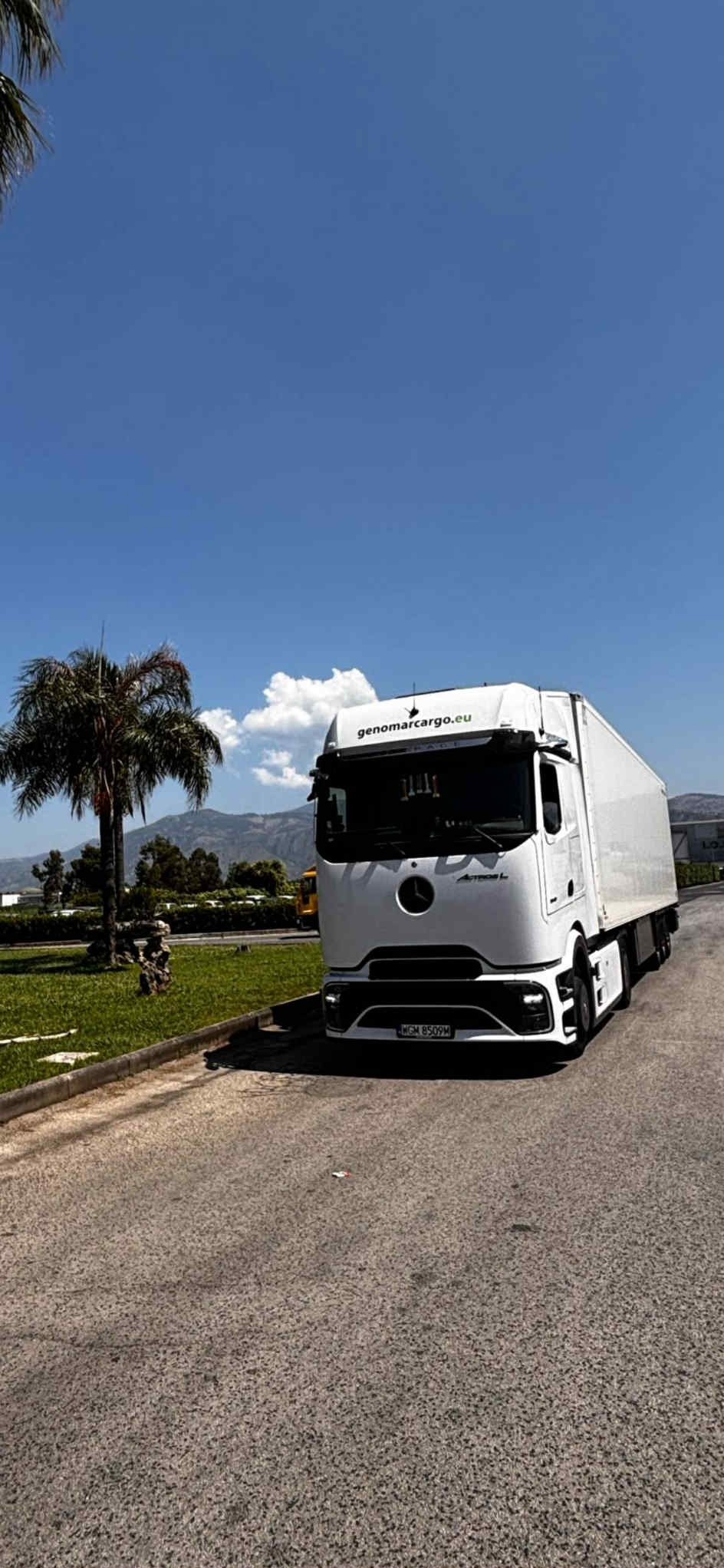 Biała ciężarówka Mercedes-Benz z naczepą na tle błękitnego nieba i gór. Na kabinie logo firmy Genomarcargo.eu. Obok palmy i zielony trawnik.
