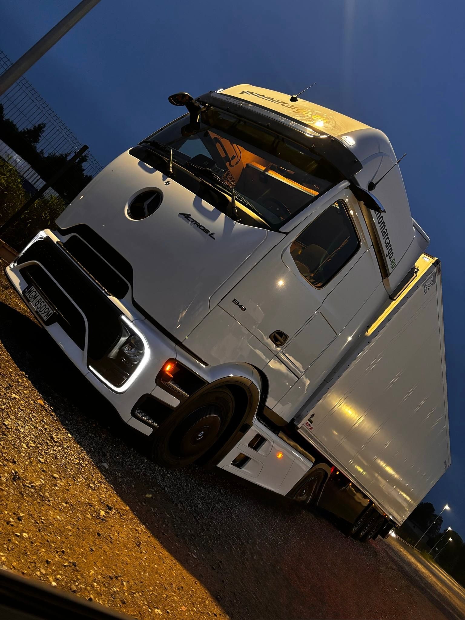 Biała ciężarówka Mercedes-Benz Actros L z naczepą, zaparkowana nocą, oświetlona światłami. Widoczne logo firmy genomarcargo.eu na dachu i boku kabiny.