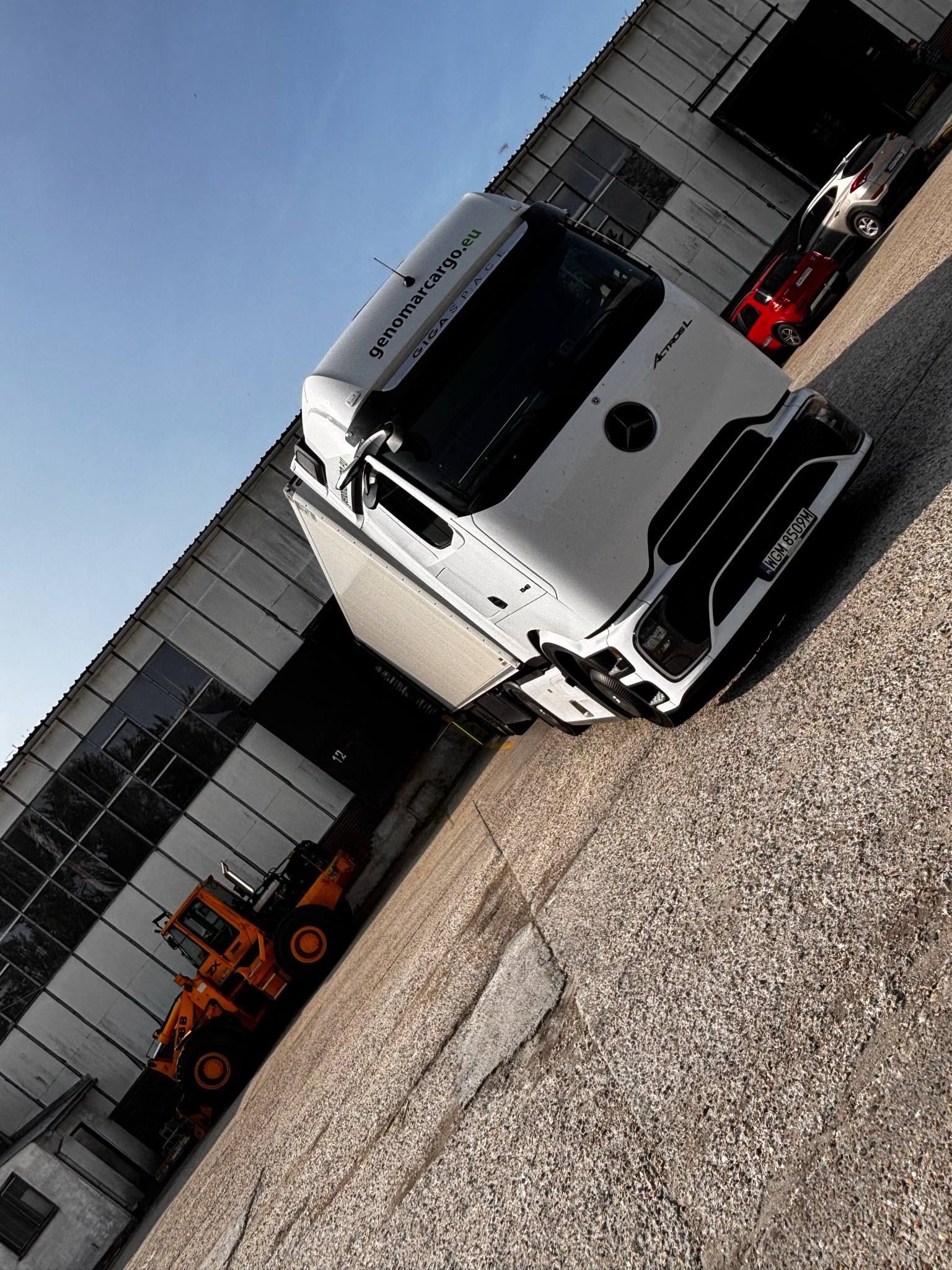 Biała ciężarówka Mercedes Actros z naczepą, zaparkowana na placu manewrowym, obok pomarańczowej ładowarki. W tle budynek magazynowy i dwa samochody osobowe.