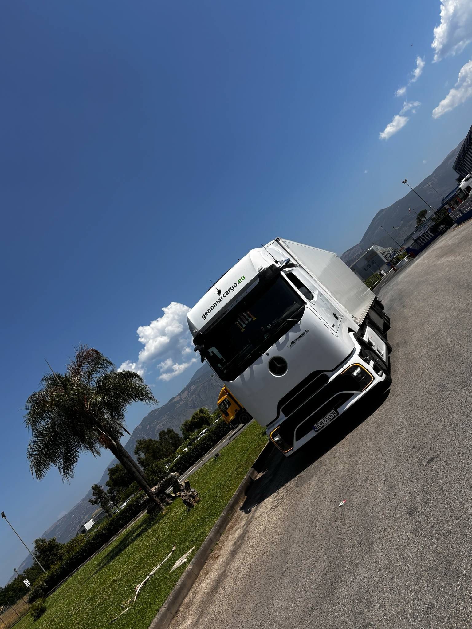 Biała ciężarówka Mercedes-Benz Actros L z logo Genomarcargo.eu na tle błękitnego nieba, gór i palmy. Widok z boku, lekko ukośny.