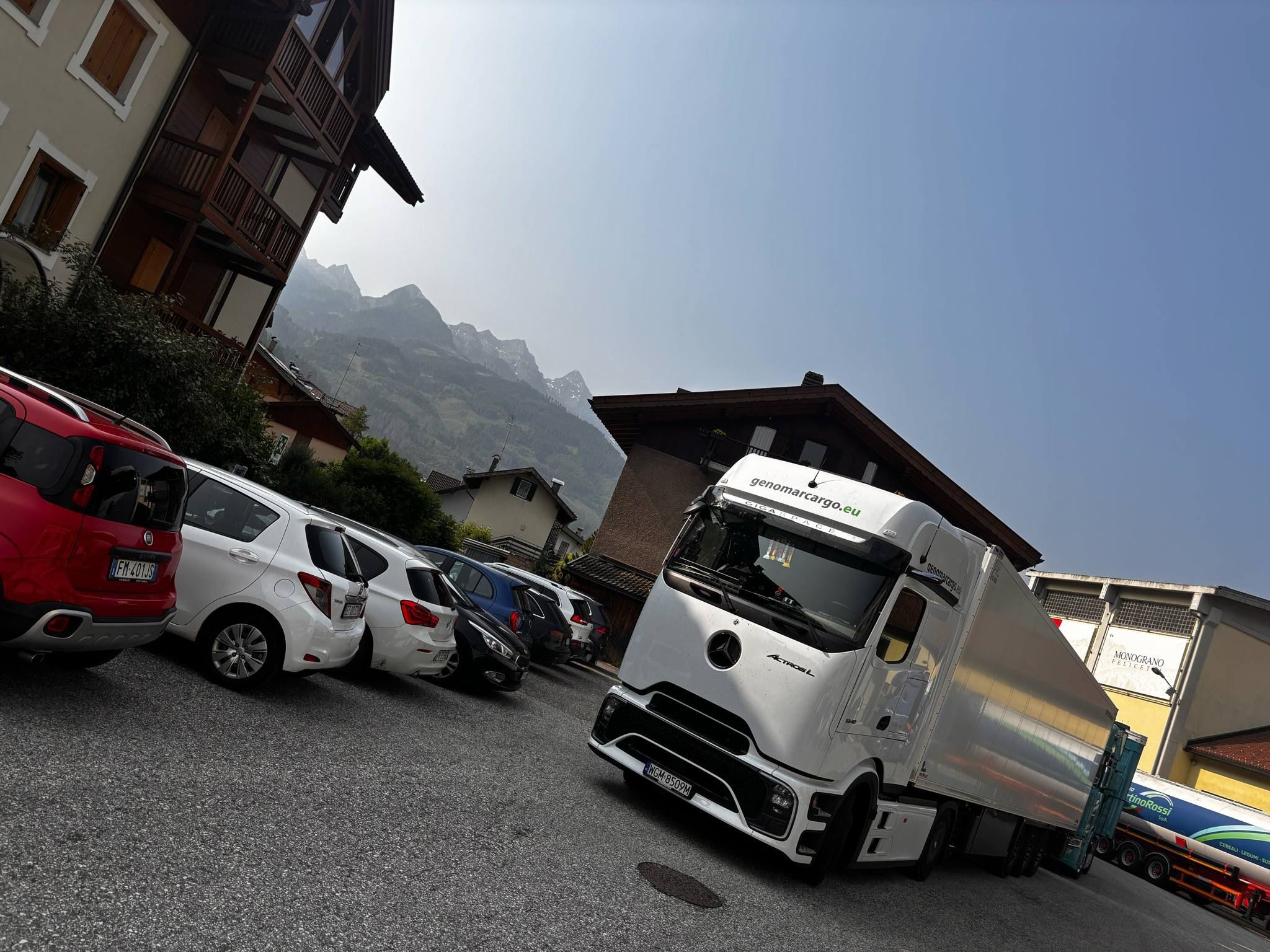 Biała ciężarówka Actros L z logo genomarcargo.eu na tle gór i parkingu z samochodami osobowymi. Ujęcie z dołu, pochylone w lewo.