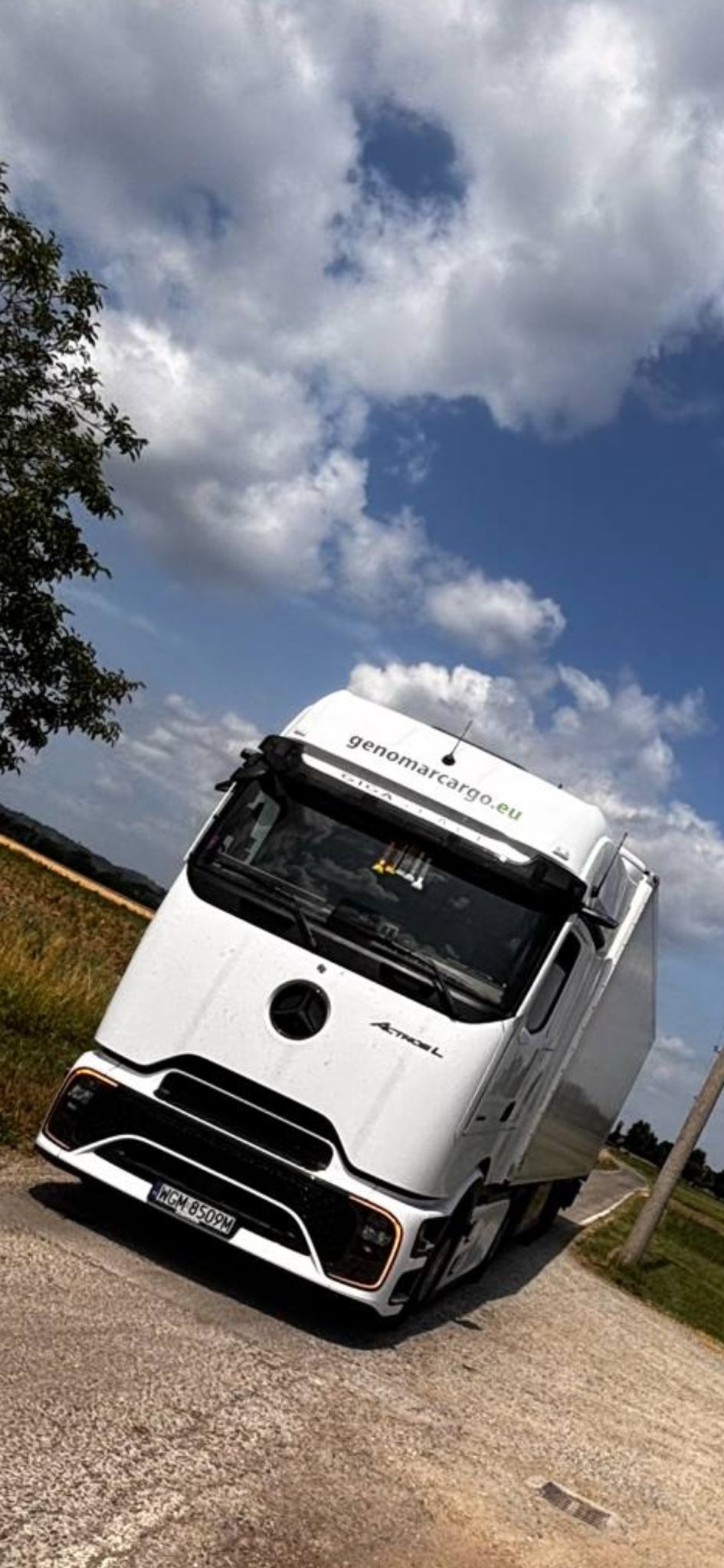 Biała ciężarówka Mercedes Actros L firmy genomarcargo.eu na wiejskiej drodze, z widocznym numerem rejestracyjnym, na tle pola i błękitnego nieba z chmurami.