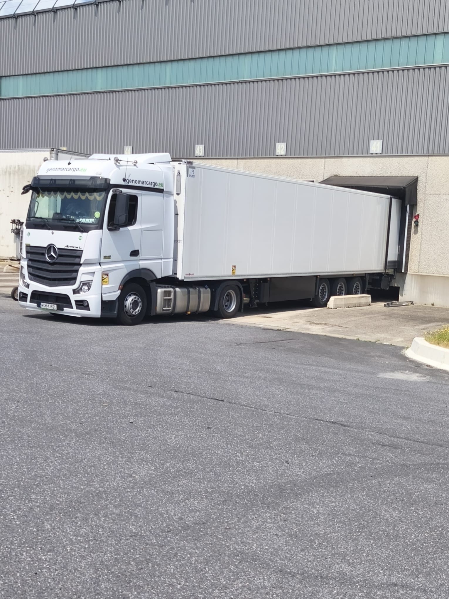 Biała ciężarówka Mercedes Actros z naczepą wjeżdża do magazynu z numerami 41-44. Na dachu widoczne logo firmy genomarcargo.eu.