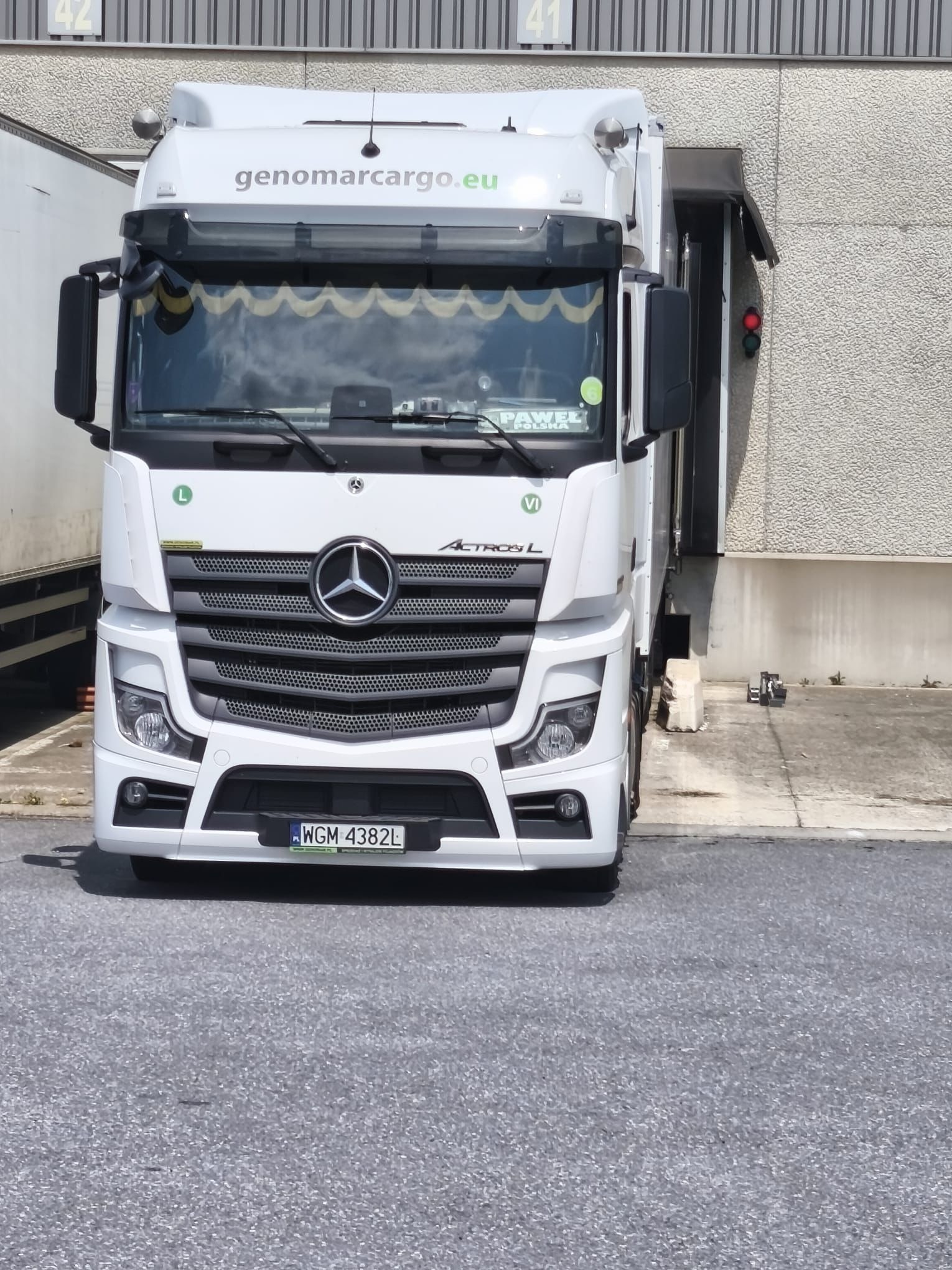 Biały ciągnik siodłowy Mercedes Actros L z naczepą, z logo genomarcargo.eu, zaparkowany pod rampą magazynową z sygnalizacją świetlną.