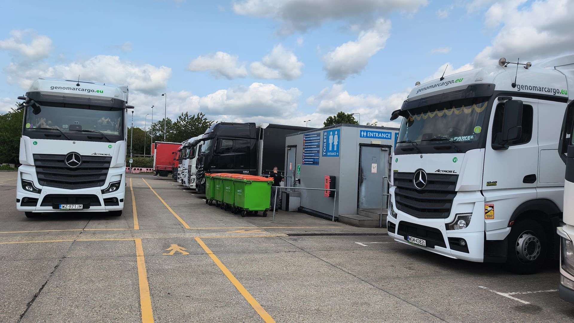 Parking dla ciężarówek z białymi Mercedesami Actros L firmy genomarcargo.eu. Na parkingu stoją zielone kontenery na śmieci. W tle widoczny budynek z toaletami.