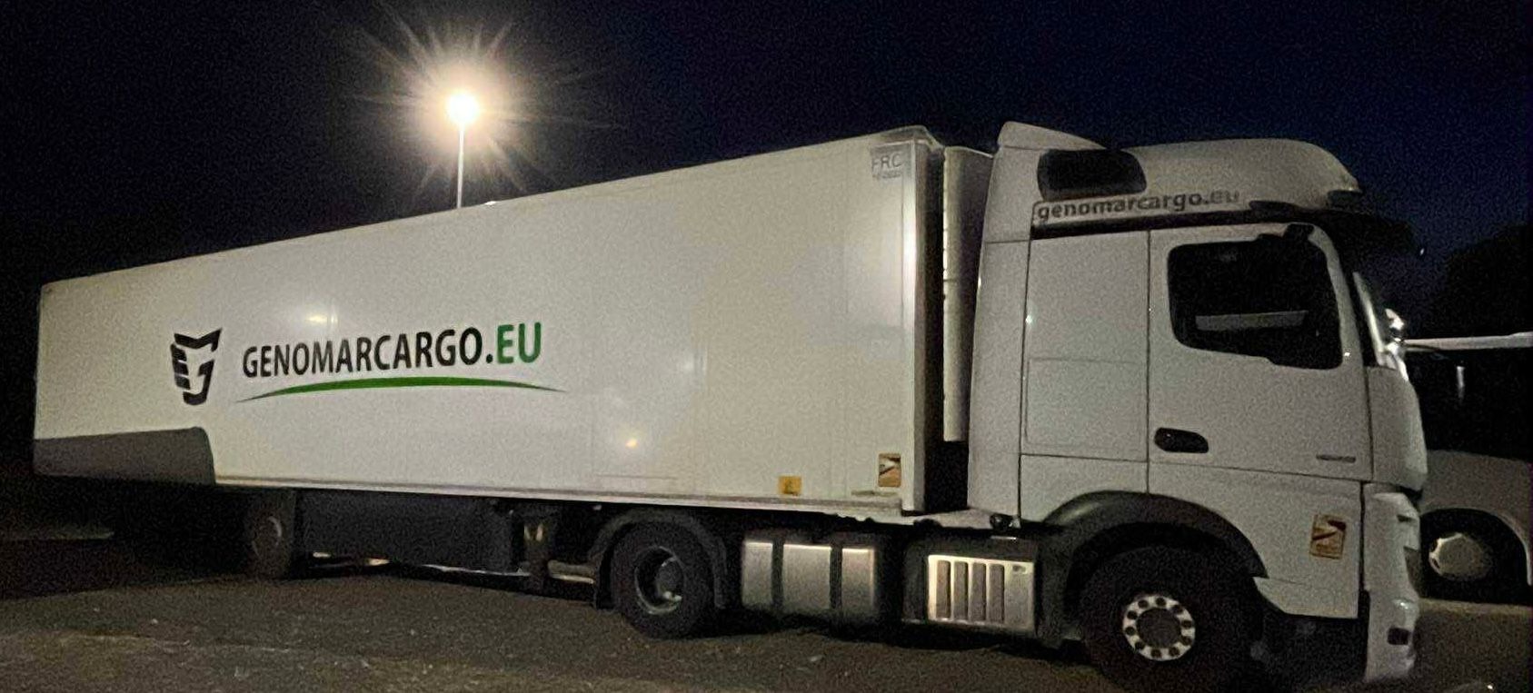 Biała ciężarówka z naczepą w nocnej scenerii, oświetlona latarnią, z widocznym logo firmy GenomarCargo.eu na boku naczepy.