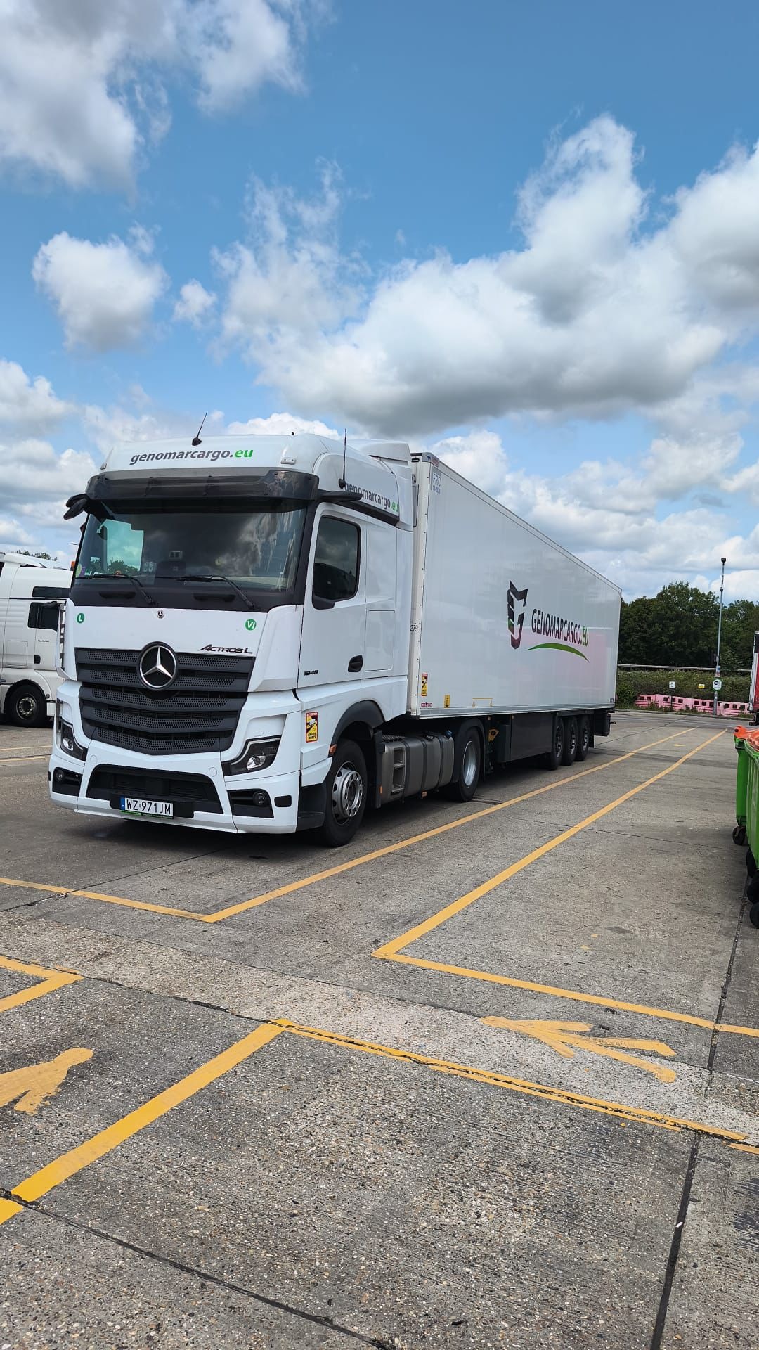 Biała ciężarówka Mercedes Actros L z logo Genomarcargo.eu na naczepie, zaparkowana na betonowym placu z żółtymi liniami. Błękitne niebo z chmurami w tle.