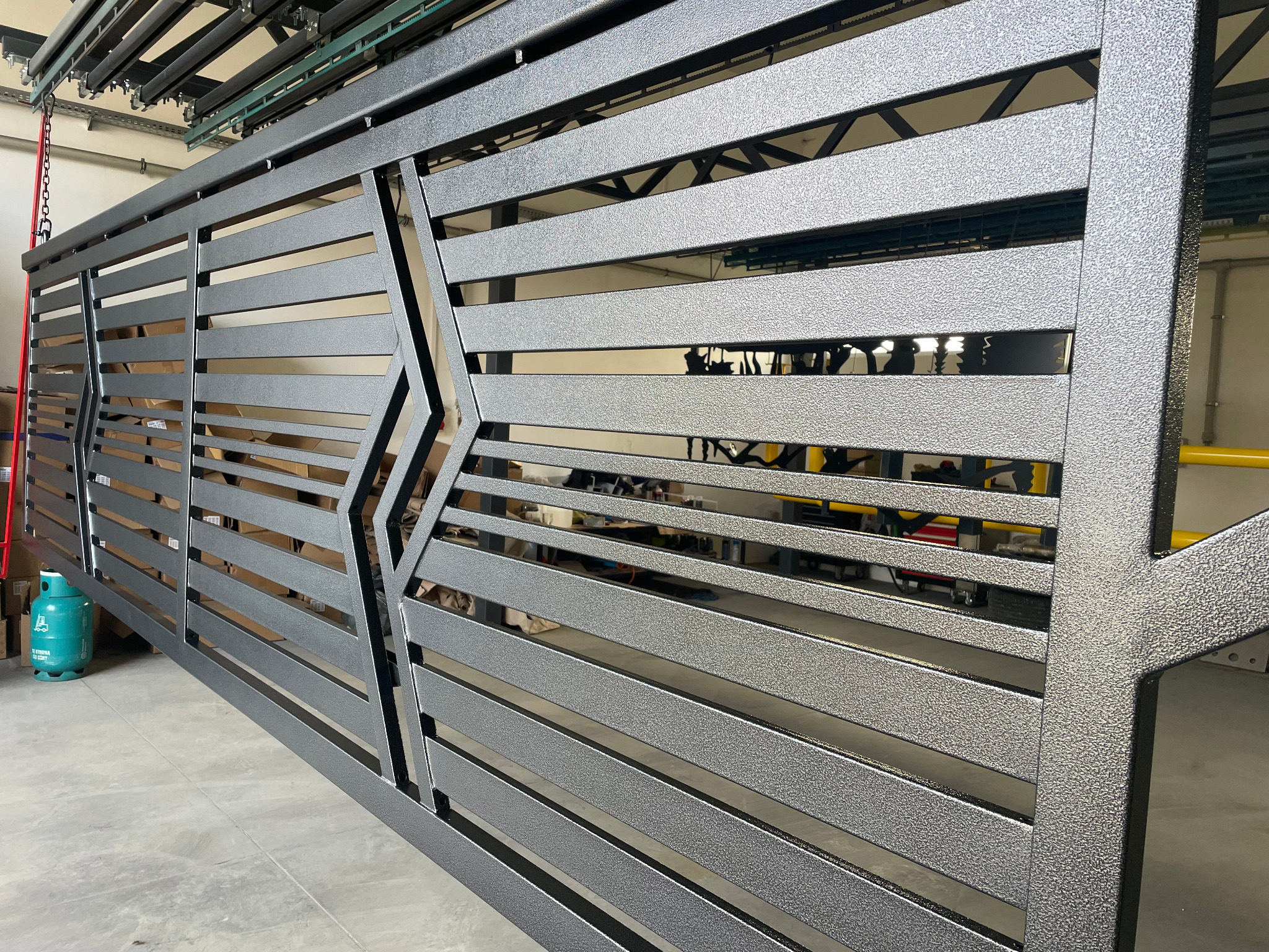 Metalowa brama przesuwna z poziomymi lamelami i geometrycznym wzorem, pokryta szarą, strukturalną farbą. Widoczny fragment linii produkcyjnej w tle.